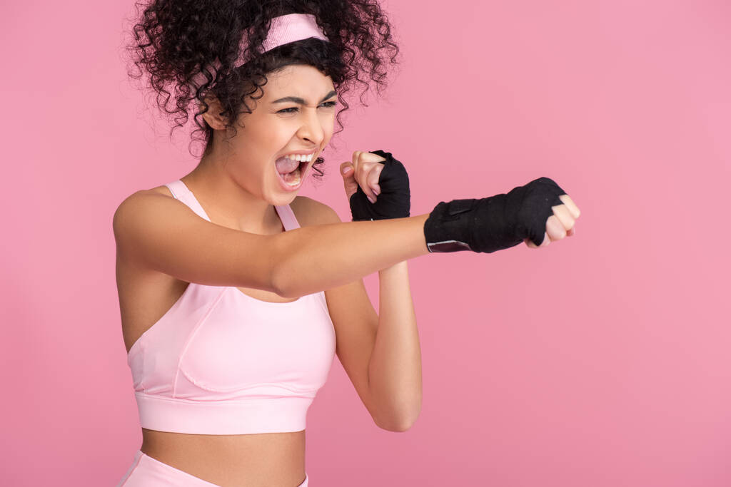 кудрявая молодая женщина в спортивной одежде с кабалой на руках упражнения изолированы на розовый - Фото, изображение