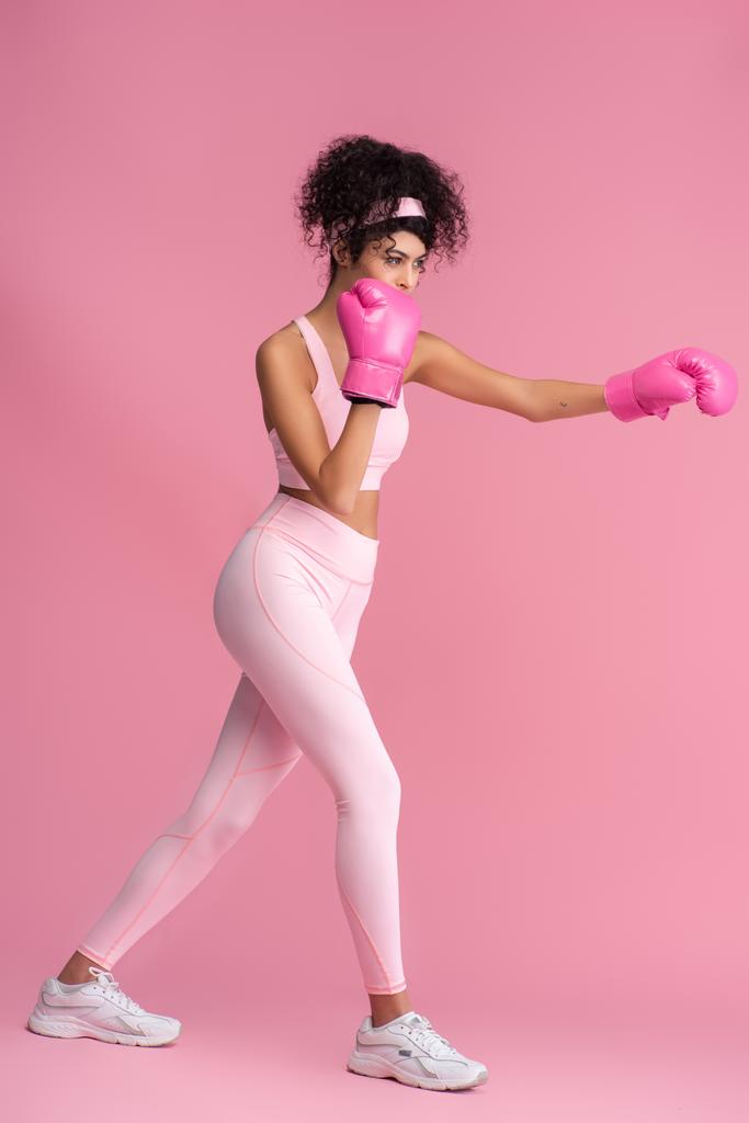 πλήρες μήκος σγουρά νεαρή γυναίκα σε αθλητικά ρούχα και γάντια πυγμαχίας εργάζονται έξω σε ροζ  - Φωτογραφία, εικόνα