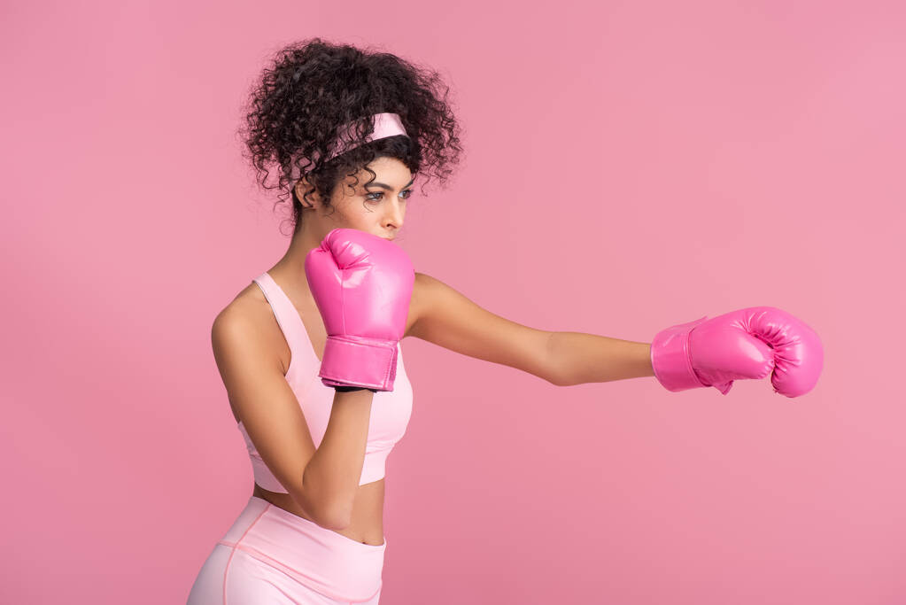 krullende jonge vrouw in sportkleding en bokshandschoenen oefenen geïsoleerd op roze  - Foto, afbeelding