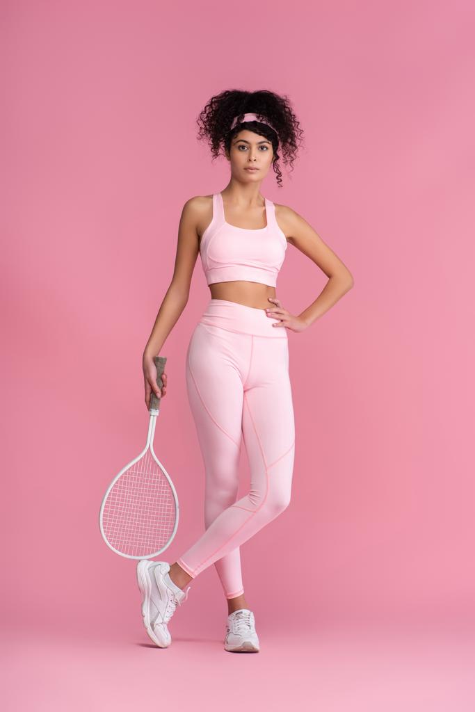 volle Länge der lockigen Frau in Sportbekleidung mit Tennisschläger im Stehen mit der Hand auf der Hüfte und gekreuzten Beinen auf rosa  - Foto, Bild