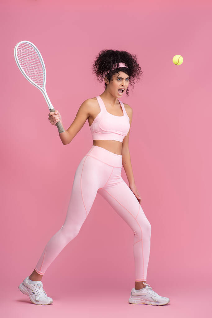 volledige lengte van krullende vrouw in sportkleding holding racket tijdens het spelen van tennis op roze  - Foto, afbeelding