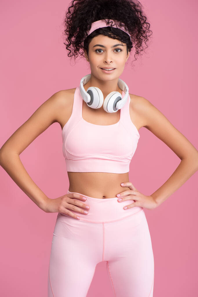 lockige junge Frau mit drahtlosen Kopfhörern, die Hände auf den Hüften isoliert auf rosa  - Foto, Bild