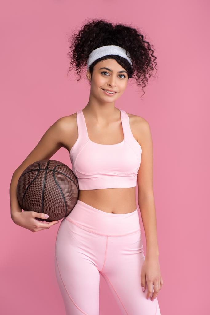 alegre joven mujer sosteniendo baloncesto aislado en rosa  - Foto, Imagen