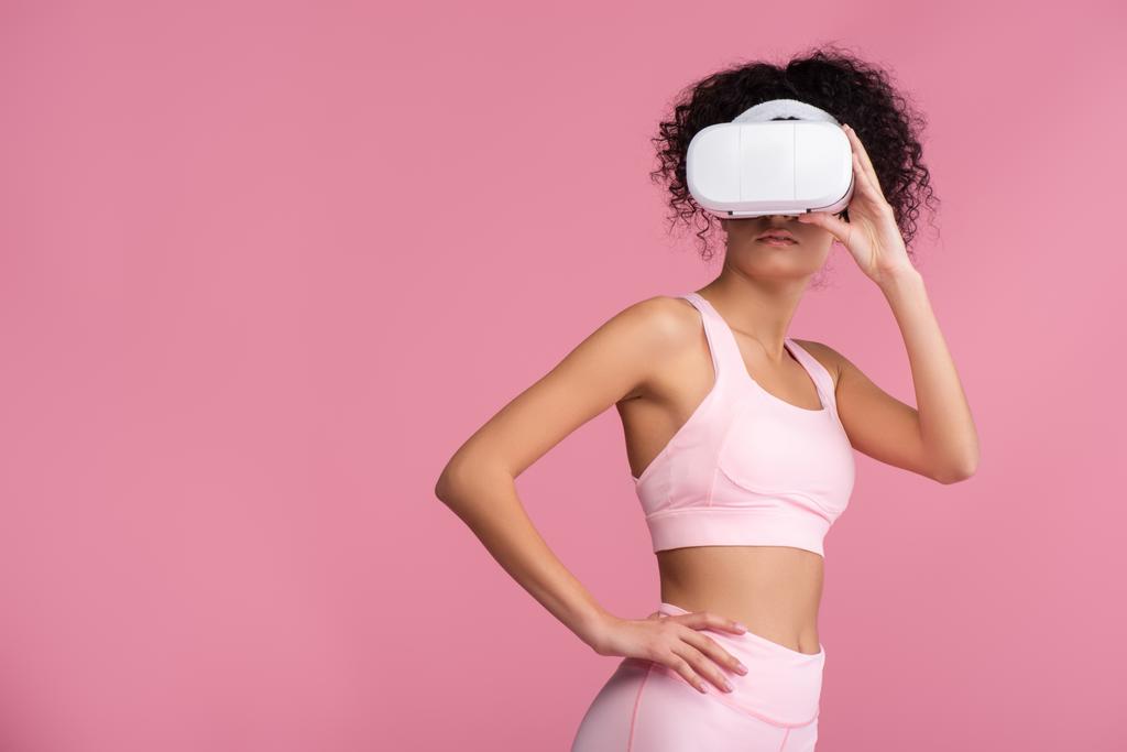 kudrnatá žena ve virtuální realitě sluchátka stojící s rukou na boku izolované na růžové - Fotografie, Obrázek