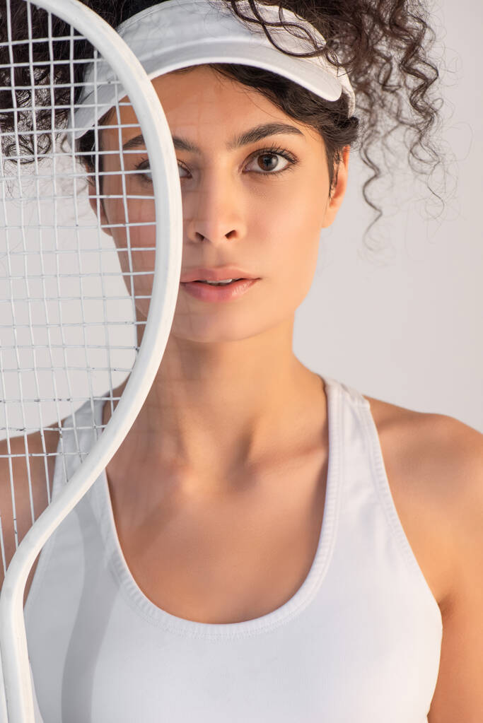 кучерява спортсменка в кепці дивиться на камеру через тенісну ракетку ізольовано на білому
 - Фото, зображення