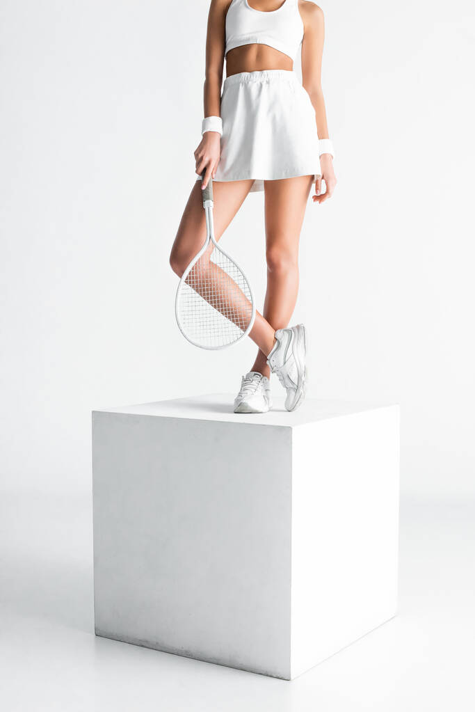 テニスラケットを持って白い立方体の上に立っている若いスポーツ女性の部分的なビュー - 写真・画像