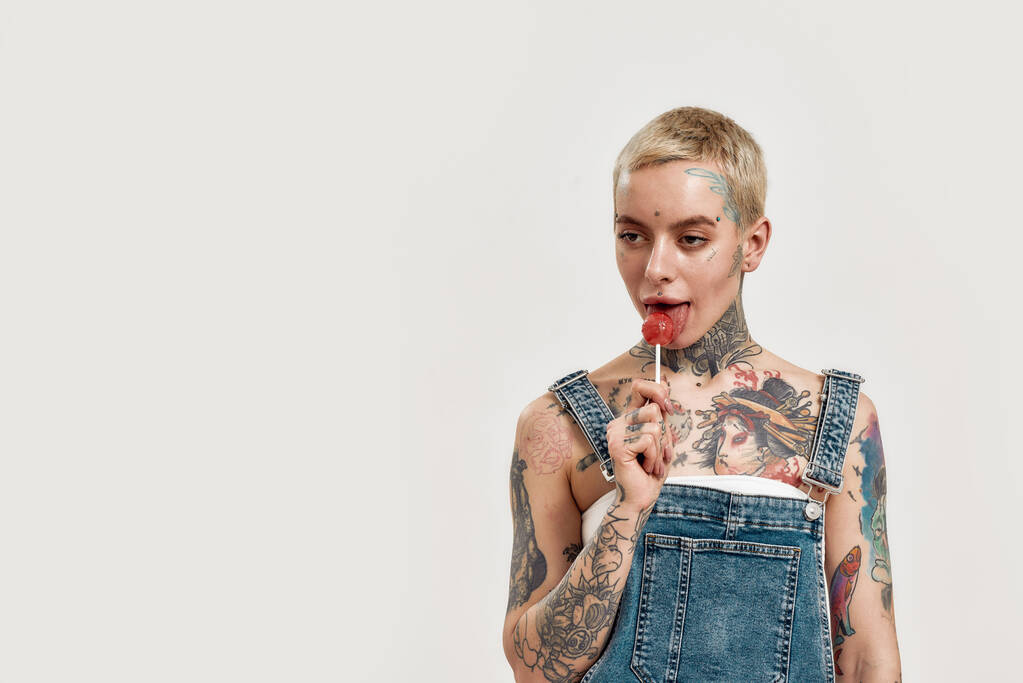 Tatuaggio e piercing. Una donna bianca traforata e tatuata che indossa un jeans lecca un lecca-lecca mentre guarda in una macchina fotografica - Foto, immagini