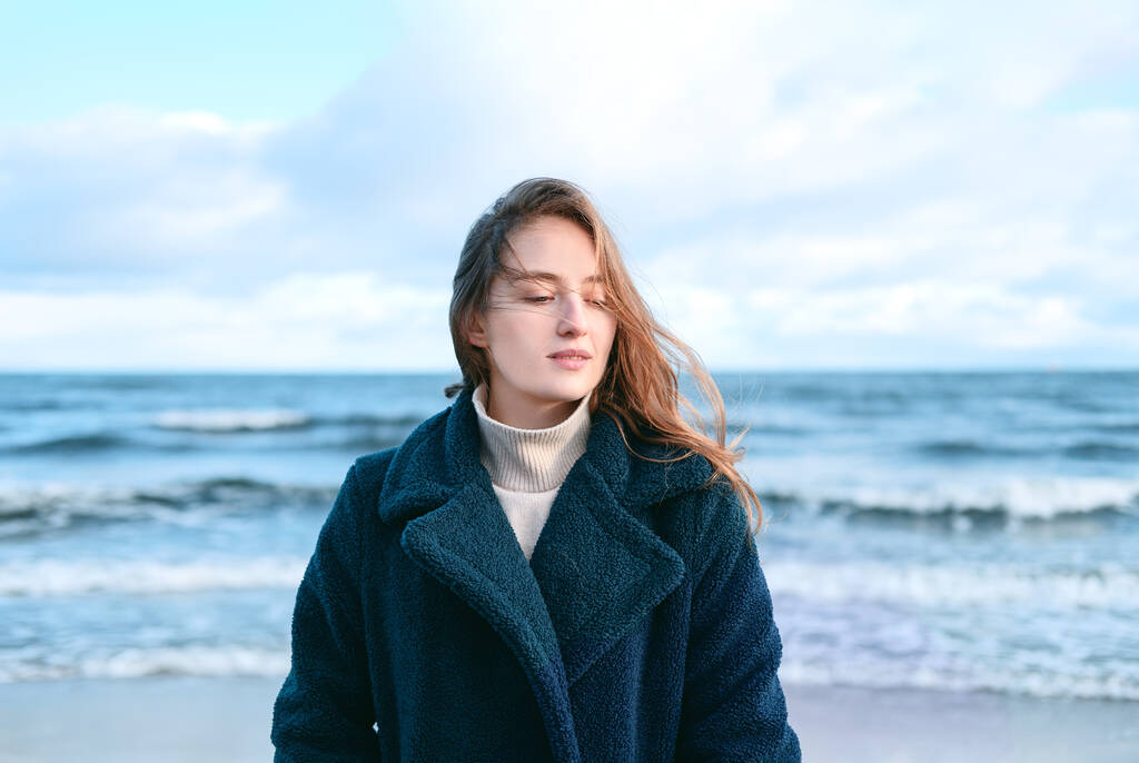 Młoda piękna kobieta walczy w mroźny zimowy dzień nad morzem. Szczęśliwa kobieta z długimi i latającymi włosami w turkusowym płaszczu odpoczywająca podczas długiej samotnej podróży stojącej na plaży i cieszącej się falą morską - Zdjęcie, obraz
