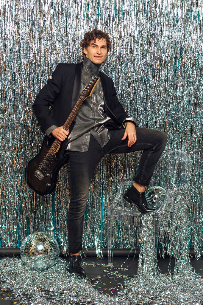 Alto uomo attraente con chitarra elettronica in piedi sopra lo sfondo scintillante con una palla da discoteca a specchio sul pavimento. Concetto di partito. - Foto, immagini