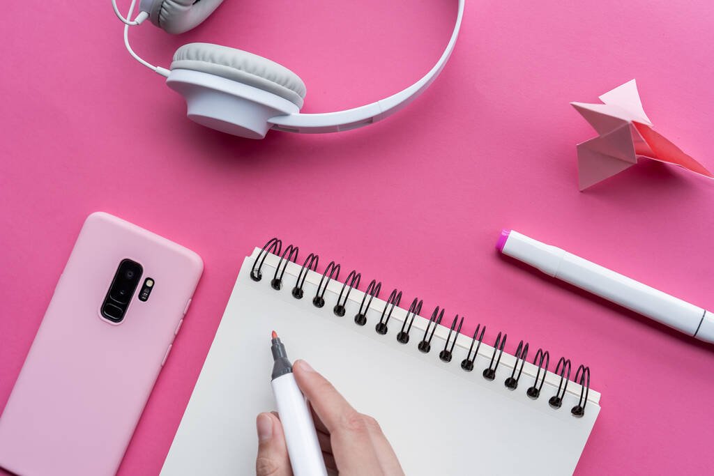 Ένα υψηλής γωνίας πλάνο ενός κινητού, ακουστικά και εργαλεία γραφής σε μια ροζ επιφάνεια - Φωτογραφία, εικόνα