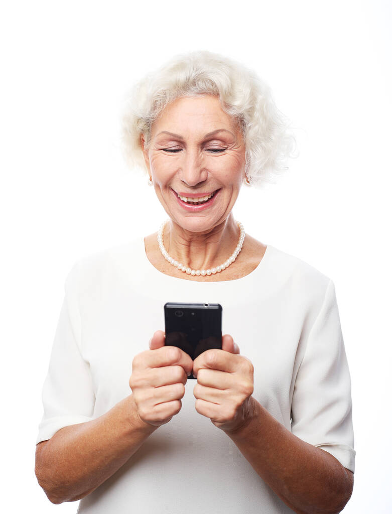 Mujer bastante mayor en traje casual usando un teléfono inteligente moderno y riendo en voz alta mientras está de pie sobre fondo blanco. Vieja viendo videos divertidos en internet. - Foto, imagen