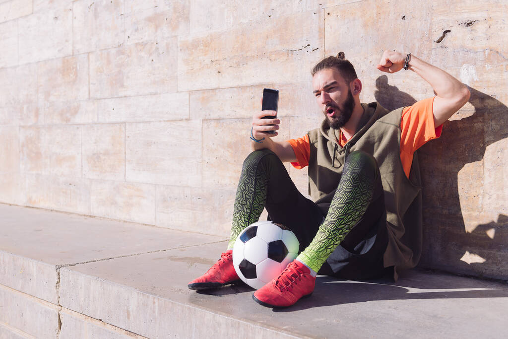 apuesto fanático del fútbol celebra una victoria cuando está consultando su teléfono inteligente, concepto de tecnología y estilo de vida deporte urbano en la ciudad, espacio de copia para el texto - Foto, Imagen