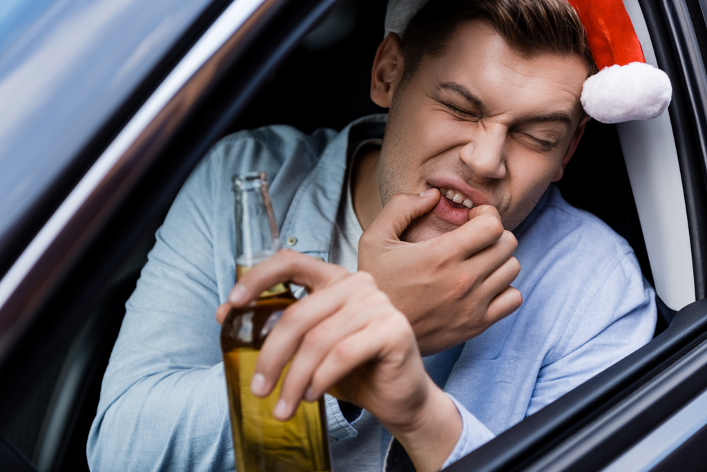 dronken man in santa hoed fluiten terwijl zitten in de auto met fles whisky, wazig voorgrond - Foto, afbeelding