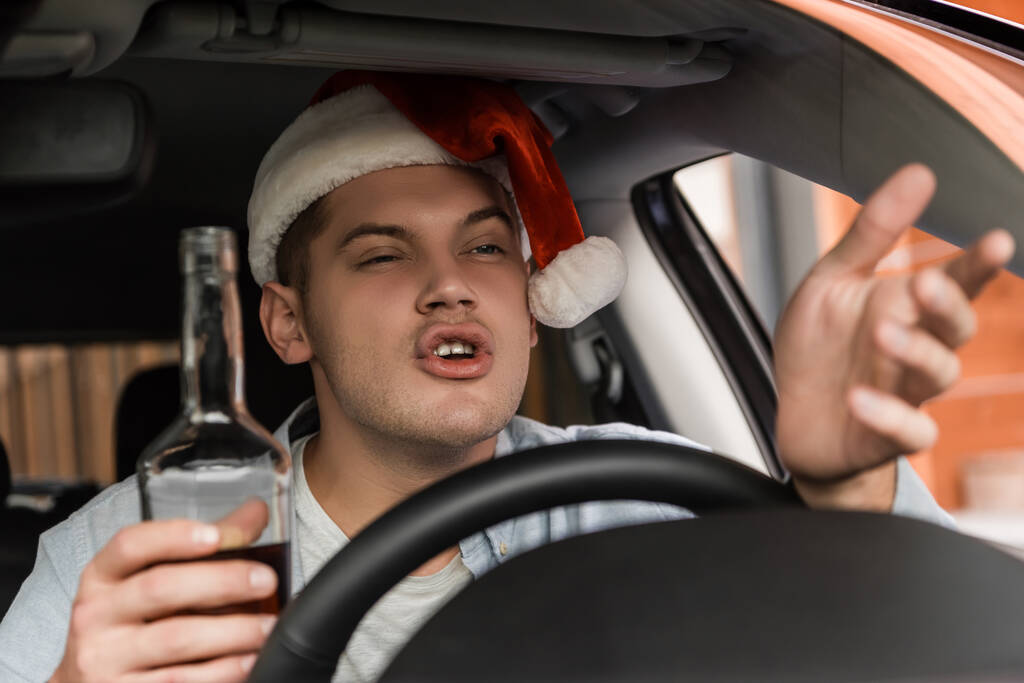 displeased, drunk man in santa hat gesturing while sitting at steering wheel with bottle of whiskey, blurred foreground - Photo, Image