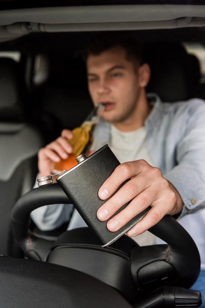 joven sosteniendo el frasco y bebiendo whisky mientras conduce el coche, fondo borroso - Foto, imagen