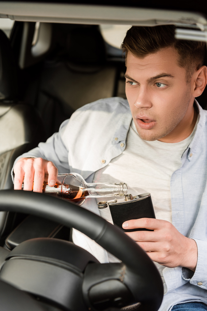 dronken man gieten whisky uit fles naar kolf tijdens het zitten in de auto, wazig voorgrond - Foto, afbeelding