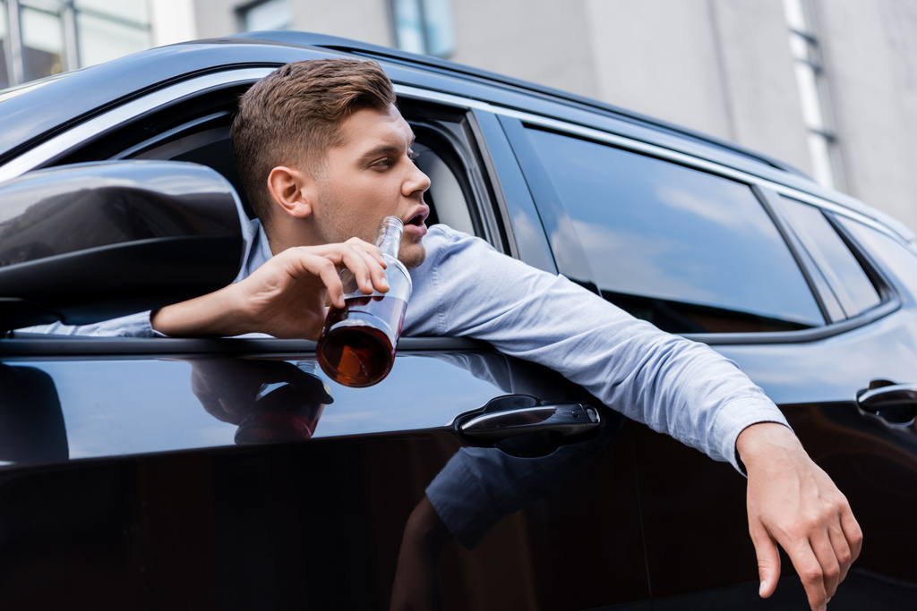 homme ivre regardant par la fenêtre de la voiture tout en tenant une bouteille de whisky - Photo, image
