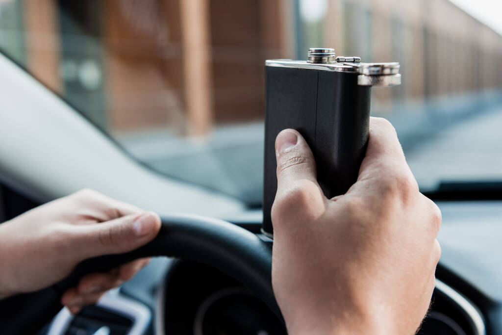 przycięty widok człowieka trzymającego kolbę z alkoholem podczas jazdy samochodem, niewyraźne tło - Zdjęcie, obraz