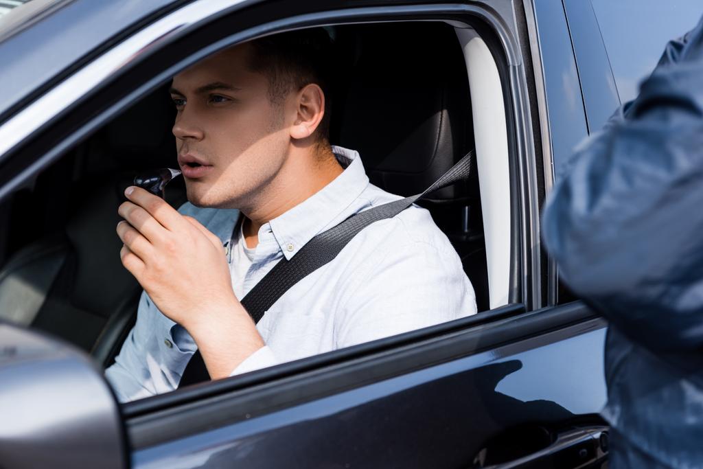 Молодой человек в машине надувает алкотестер, а полицейский стоит на размытом переднем плане - Фото, изображение