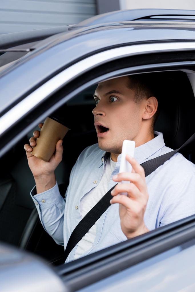 шокированный мужчина держит кофе, чтобы пойти и мобильный телефон на водительское сиденье в машине, размытый передний план - Фото, изображение