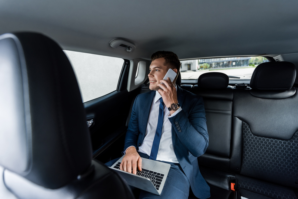Smiling businessman talking on smartphone and using laptop in car - Photo, Image