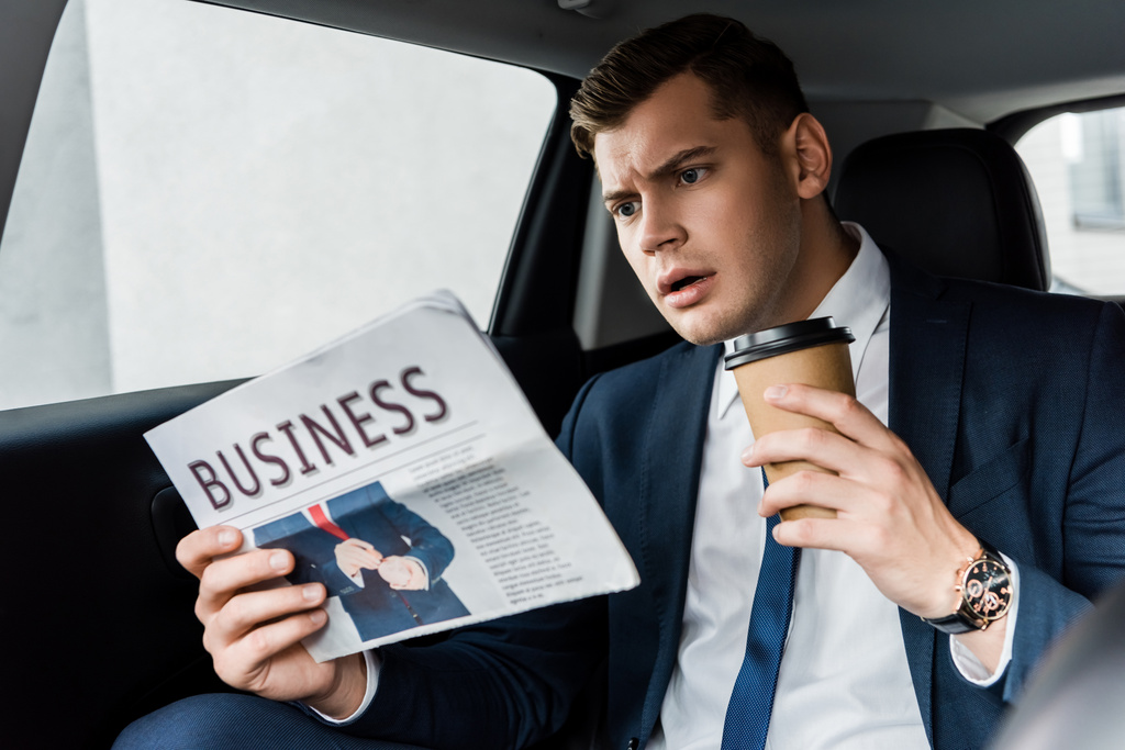 Сосредоточенный бизнесмен с одноразовой чашкой читает газету в авто - Фото, изображение