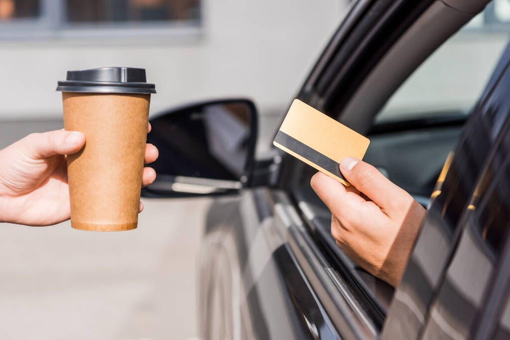 Vista recortada del vendedor sosteniendo café para acercarse al hombre con tarjeta de crédito en auto - Foto, imagen