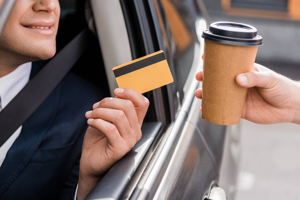 Przycięty widok uśmiechniętego biznesmena w samochodzie z kartą kredytową blisko sprzedawcy z kawą na wynos - Zdjęcie, obraz