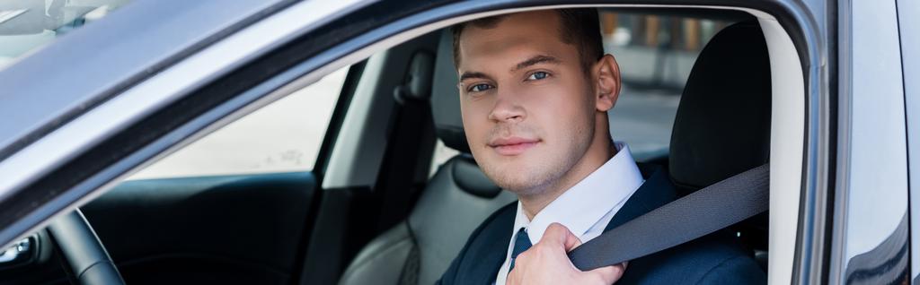 Nuori liikemies katselee kameraa pitäen turvavyötä autossa hämärtyneellä etualalla, banneri - Valokuva, kuva