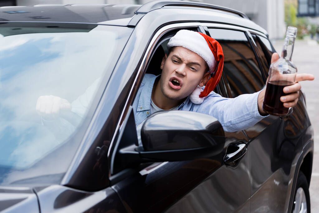 Betrunkener, wütender Mann mit Weihnachtsmütze, mit Flasche Whiskey, der während der Autofahrt aus dem Fenster schaut - Foto, Bild