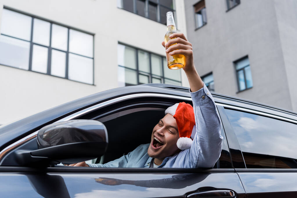 excitado, hombre borracho en sombrero de santa sosteniendo botella de alcohol en la mano levantada mientras está sentado en el coche - Foto, Imagen