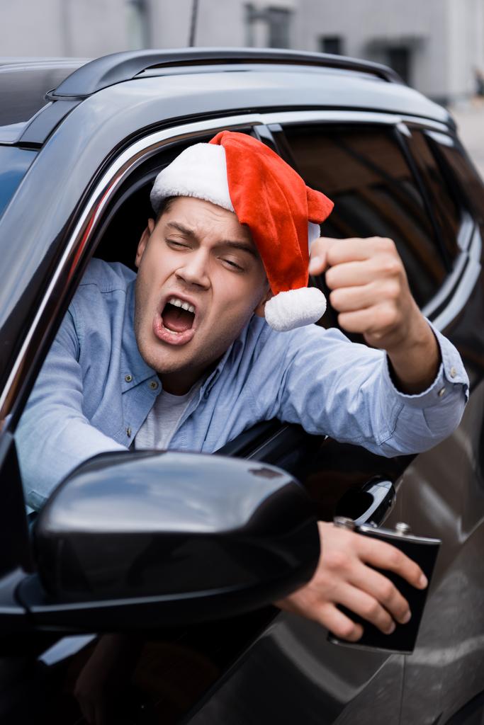 aggressiver, betrunkener Mann mit Weihnachtsmannmütze, mit Alkoholflasche, geballte Faust beim Blick aus dem Autofenster, verschwommener Vordergrund - Foto, Bild