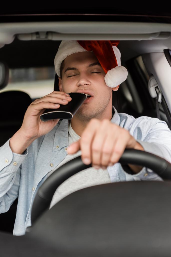 Betrunkener mit Weihnachtsmannmütze trinkt mit geschlossenen Augen aus Kolben, während er Auto auf verschwommenem Vordergrund fährt - Foto, Bild