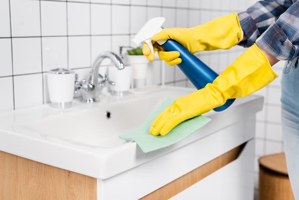 Vista recortada de mujer joven en guantes de goma fregadero de limpieza con detergente y trapo en el baño  - Foto, imagen