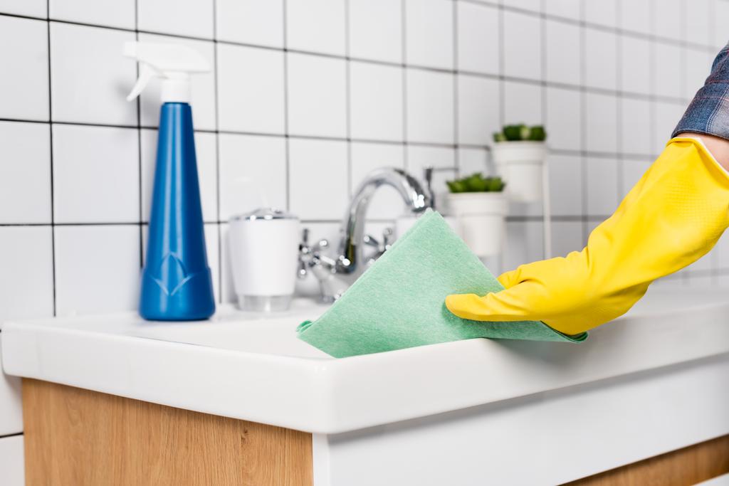 Lastik eldivenli bir kadının banyo lavabosunu temizlerken çekilmiş görüntüsü bulanık arka planda deterjan şişesinin yanında.  - Fotoğraf, Görsel