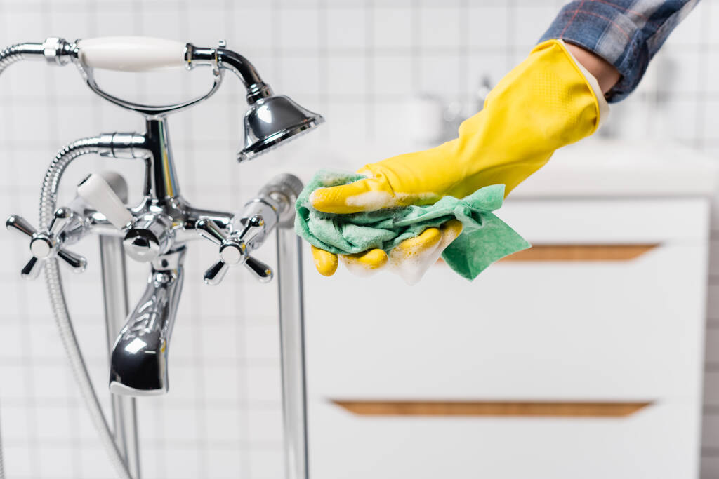 Gewassen uitzicht van de vrouw in rubber handschoen houden doek met zeep in de buurt kraan en douche in de badkamer  - Foto, afbeelding
