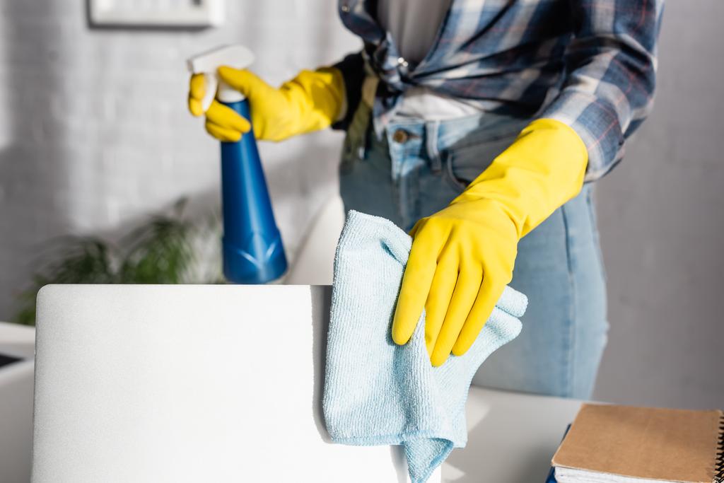 Cropped view of woman in rubber gloves cleaning laptop with rag and holding detergent on blurred background  - Photo, Image