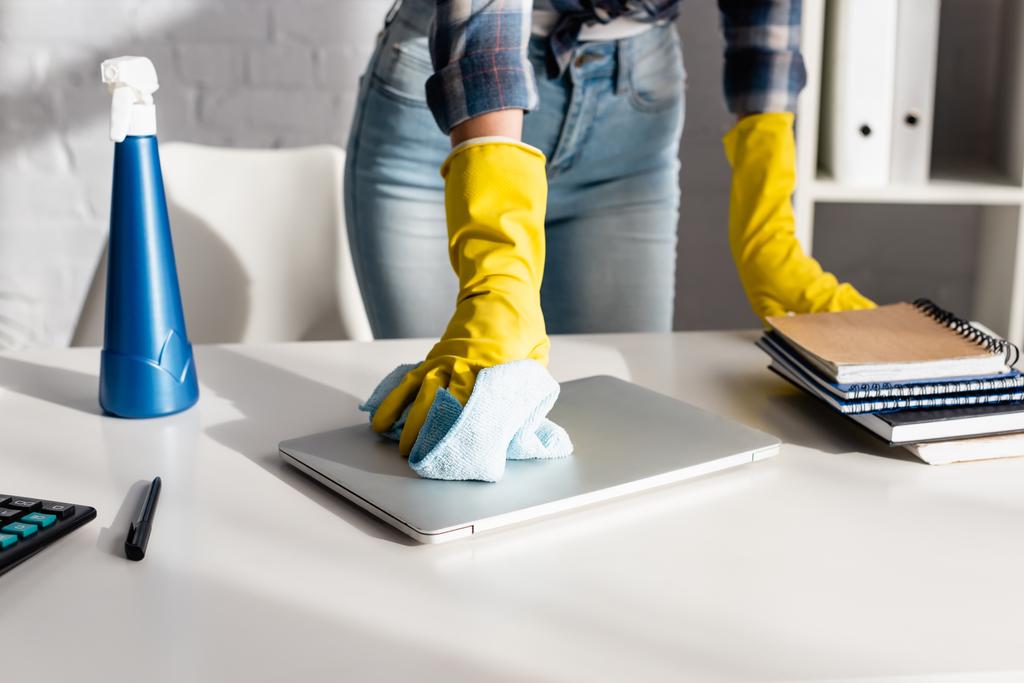Vista recortada de la mujer en guantes de goma limpieza portátil cerca de detergente y cuadernos sobre fondo borroso en la mesa  - Foto, imagen
