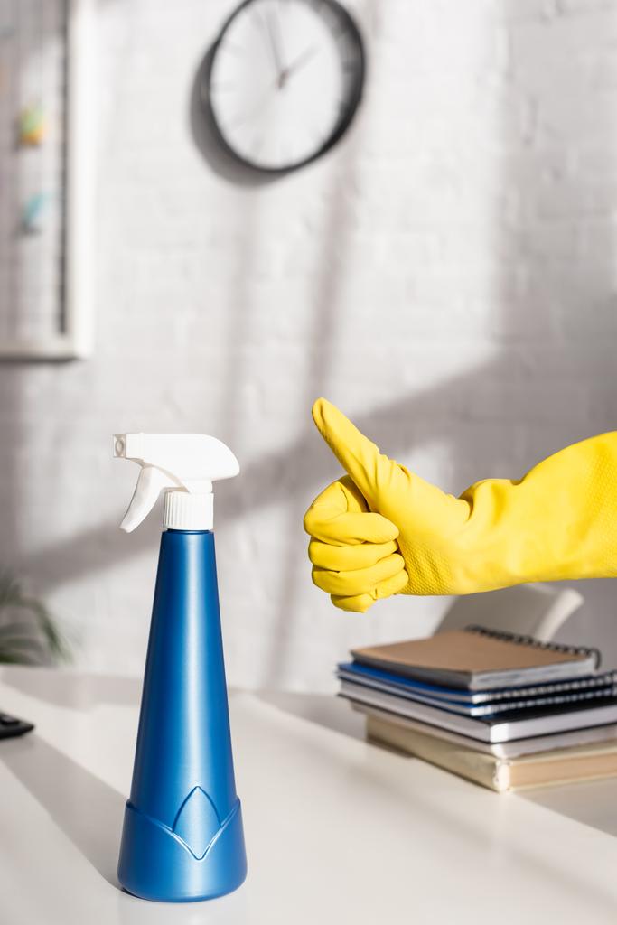 Vista recortada de la botella de detergente y mano en guante de goma mostrando como gesto cerca de la mesa  - Foto, imagen