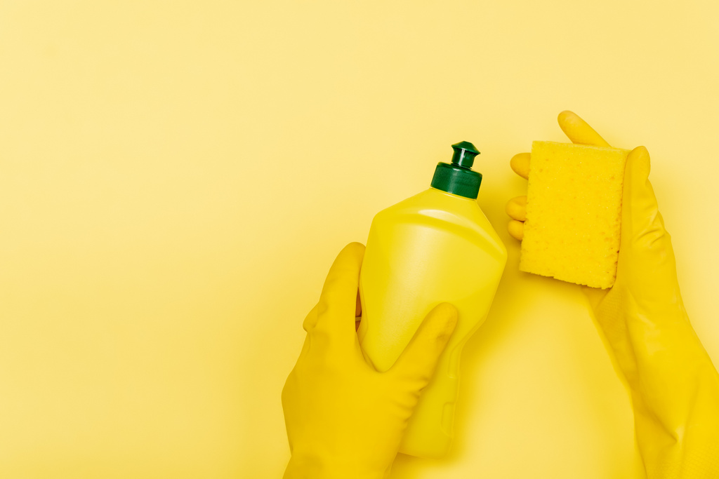 Draufsicht der Hände in Gummihandschuhen mit einer Flasche Spülmittel und Schwamm auf gelbem Hintergrund - Foto, Bild