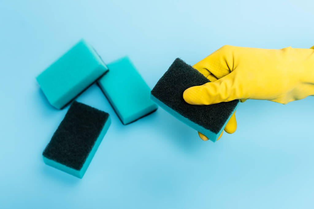 Ansicht der Hand in Gummihandschuhen mit Schwamm auf blauem Hintergrund - Foto, Bild