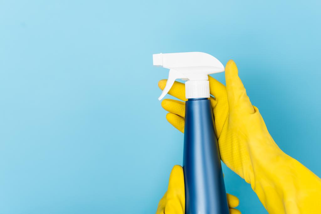 Vista ritagliata delle mani in guanti di gomma gialla tenendo detergente con spruzzatore su sfondo blu - Foto, immagini