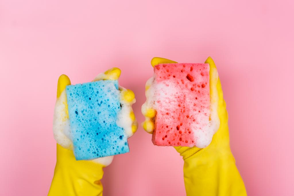 Vista superior de las manos en guantes de goma sosteniendo esponjas con espuma de jabón sobre fondo rosa - Foto, Imagen