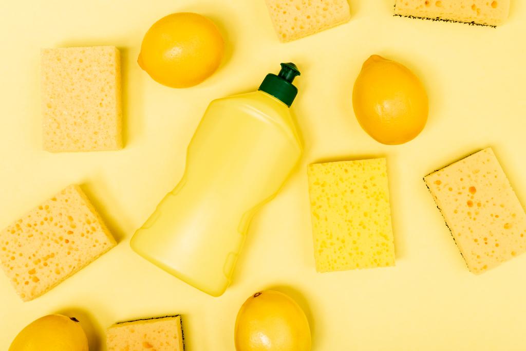 Вид сверху целых лимонов рядом с губками и жидкостью для мытья посуды на желтом фоне - Фото, изображение