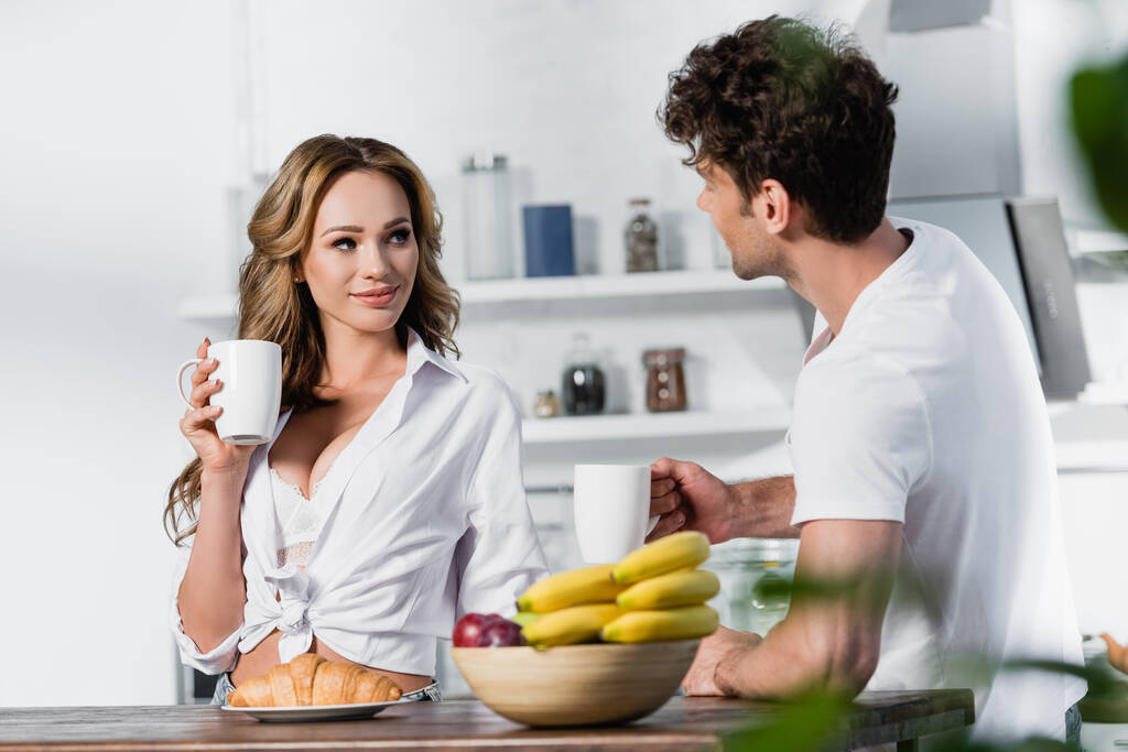 Mujer sexy sosteniendo taza cerca del desayuno y novio en primer plano borrosa en la cocina  - Foto, Imagen