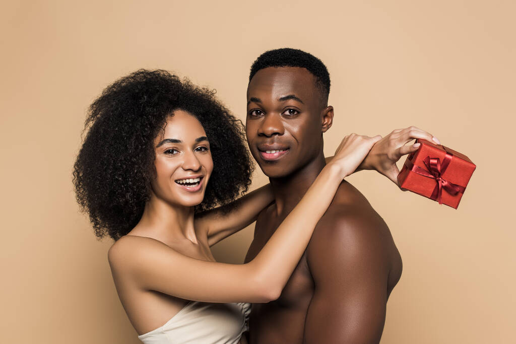 happy african american woman holding present and hugging shirtless boyfriend isolated on beige - Photo, Image