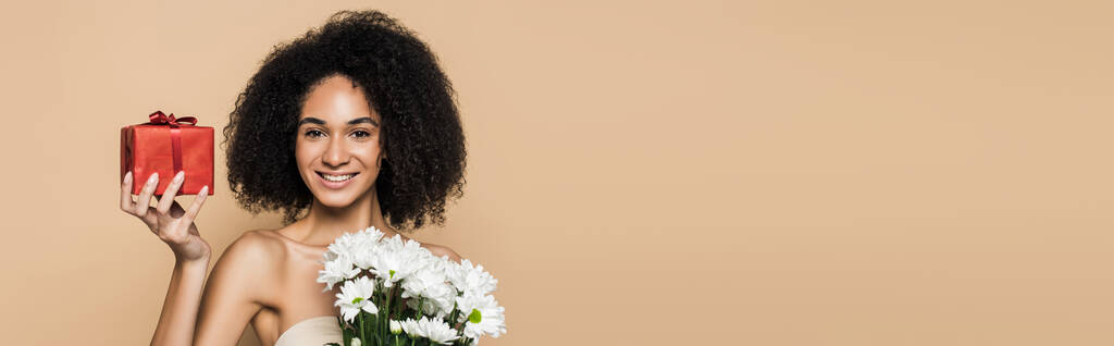 весёлая африканская американка с голыми плечами, держащая в руках подарок и цветы, изолированные на бежевом, баннере - Фото, изображение