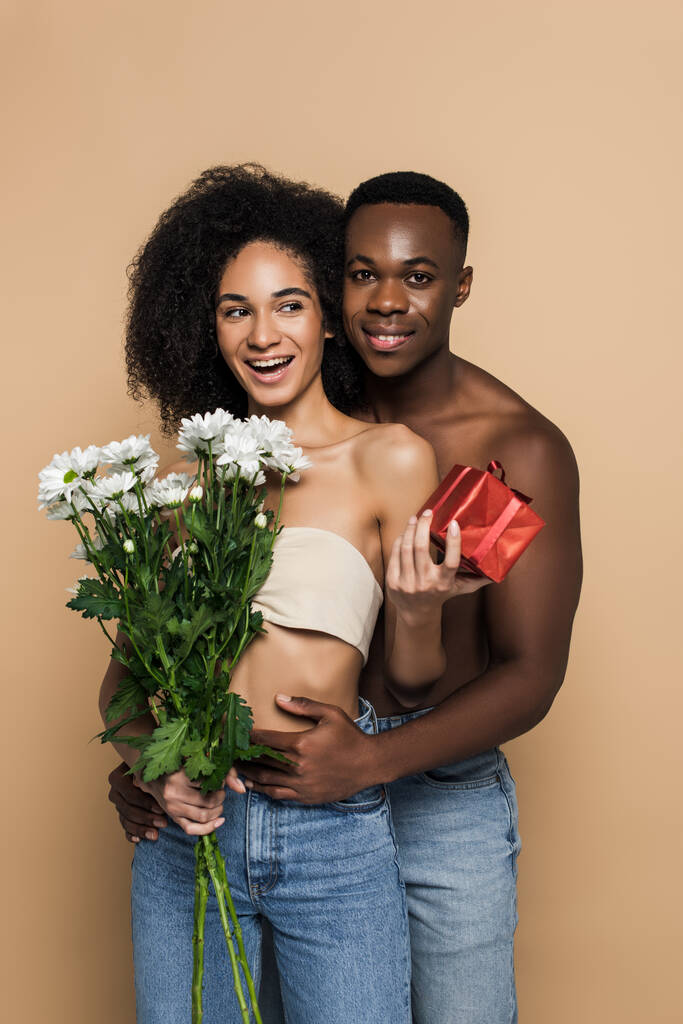 Üstsüz, Afro-Amerikalı bir adam. Kıvırcık bir kadını çiçeklerle kucaklıyor ve bej rengi bir elbiseyle sunuyor. - Fotoğraf, Görsel