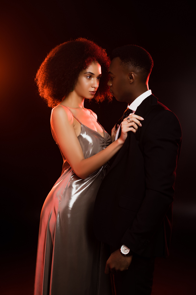 curly african american woman in dress talking off blazer on boyfriend in suit on black - Photo, Image