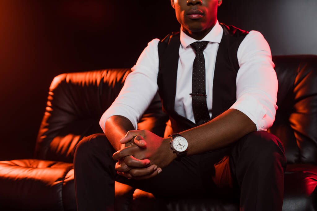 vista recortada del hombre afroamericano en traje sentado en el sofá con las manos apretadas sobre fondo negro  - Foto, Imagen